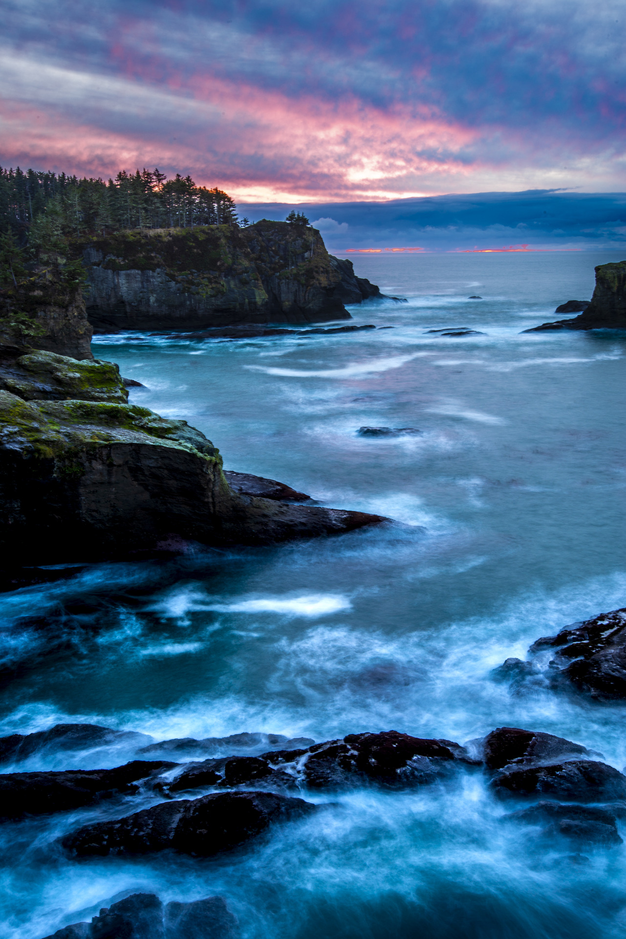 Long exposure of Cape Flattery coastline at sunset — flowing water, dark cliffs, and fire clouds